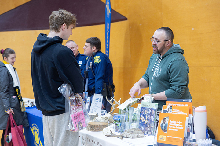 A male student standing next to a table at MainStage while with a man stands behind he table