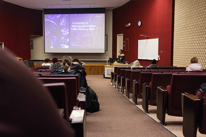 A wide angle shot of an auditorium. On a screen is a presentation titled A Century of Misrepresentation.
