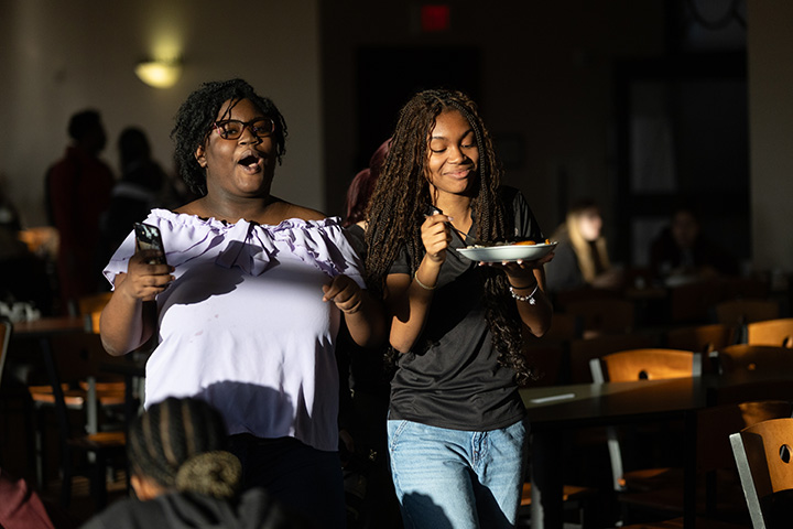 Two young women standing up, holding a plate and a fork.