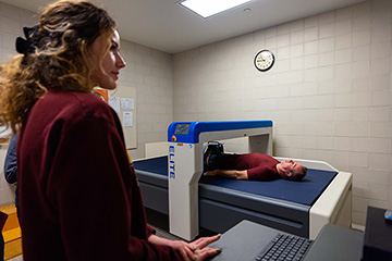 CMU President MacKinnon lying in a DEXA scan machine while two people run a computer