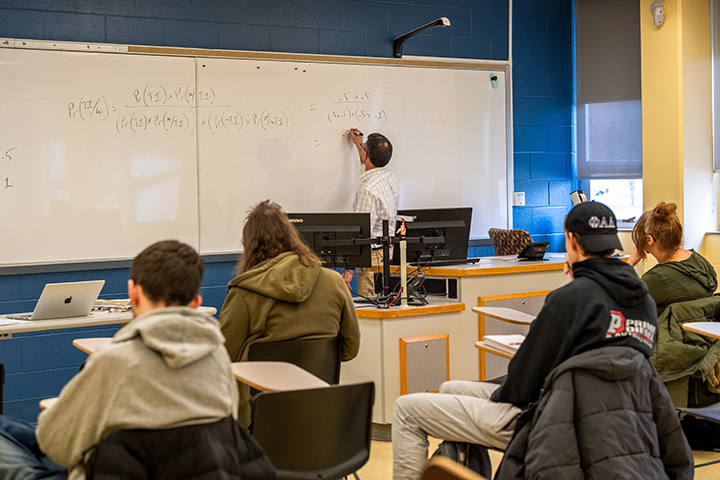 A professor writing on a whiteboard in a classroom full of students