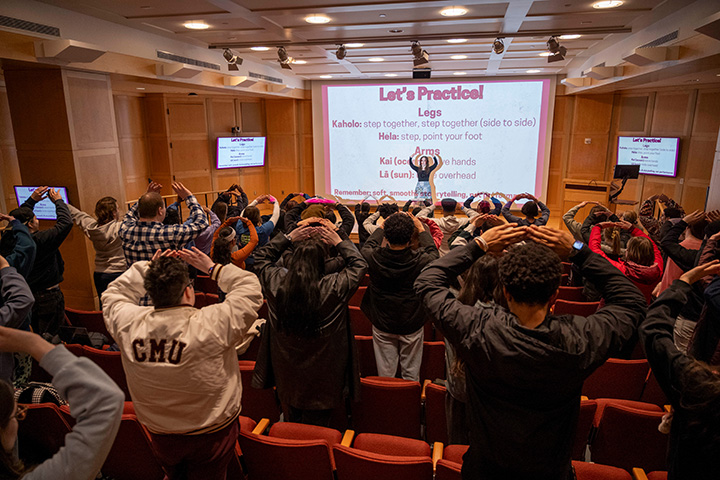 A group of people in an auditorium put their hands in the air while learning to do the hula