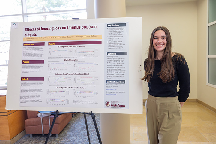 A student smiles for the camera while standing next to a display board containing research details