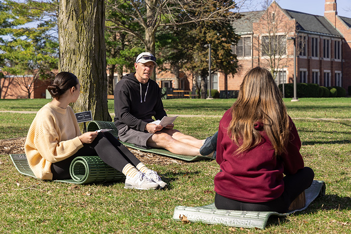 Three people sit on yoga mats in the grass underneath a shady tree