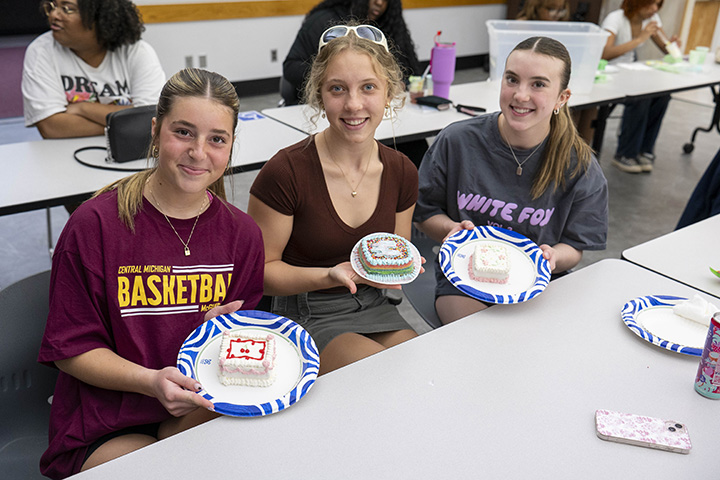 Three students hold plates with pieces of cake on them while sitting at a table in a classroom