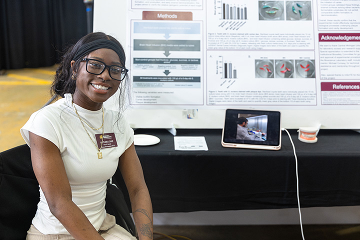 A student sits in a chair next to a table with a poster board showing research results