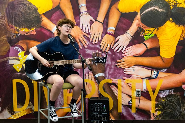 A CMU student plays a guitar and sings while sitting on a tall chair