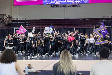 A large group of student fraternity and sorority members all dressed in black dance on the court at CMU's McGuirk Arena