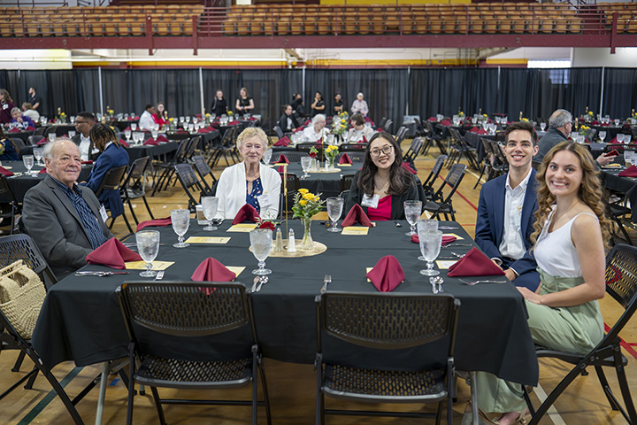 A group of two elderly people and three students sit at a large square table for a dinner reception inside Finch Fieldhouse