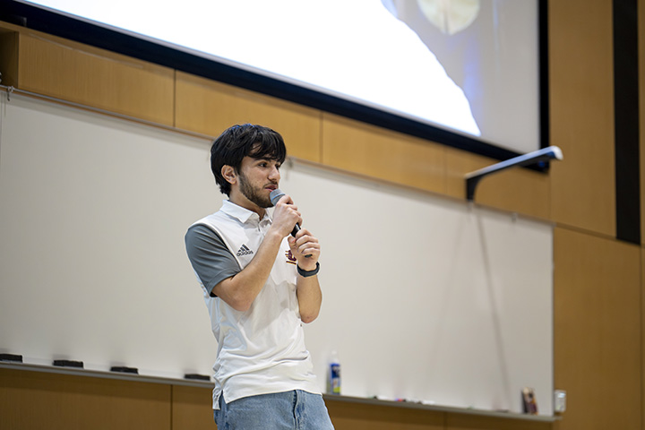 A bearded male student stands in front of a classroom holding a microphone
