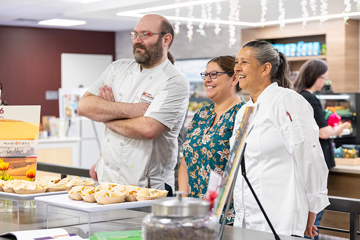 Three people, two in chef jackets, stand near a table that has food on it