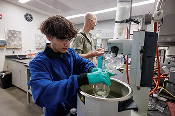 Inside a research lab, a student in a blue jacket holds a beaker over a bucket