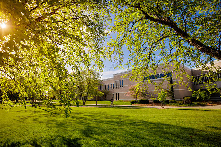 Looking through two trees at the Music Building as the sun pokes through the branches and a student walks down a sidewalk