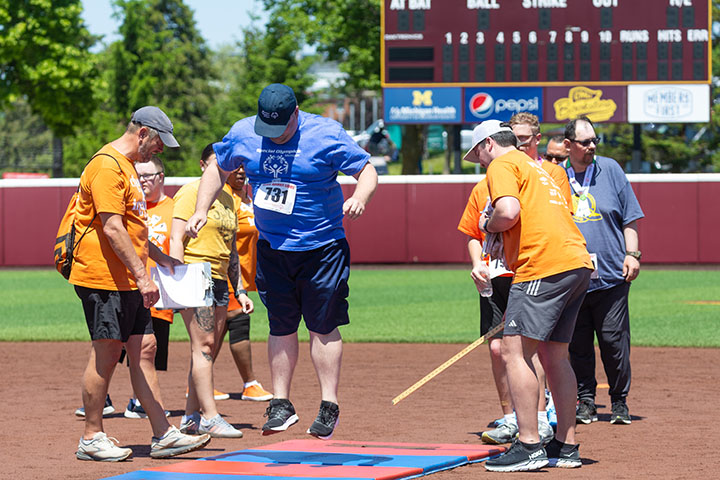 An athlete competes in standing long jump while volunteers in orange shirts prepare to measure the jumping distance