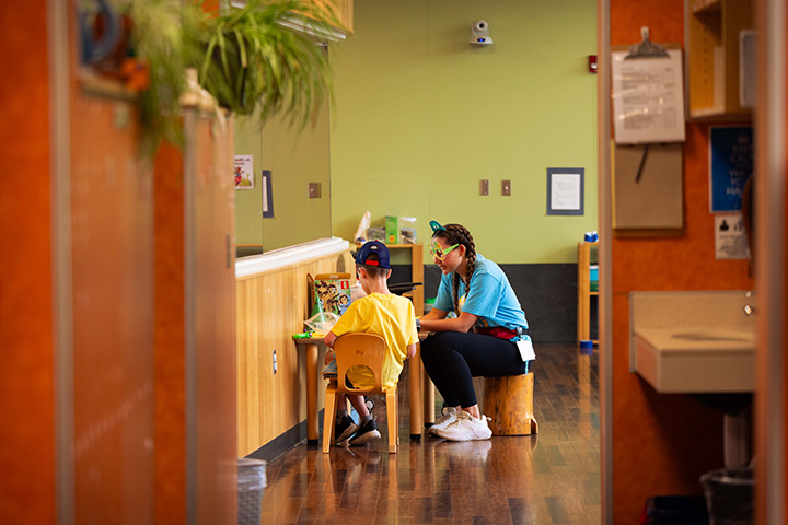 Looking through a doorway, an elementary student sits at a small table with a teacher