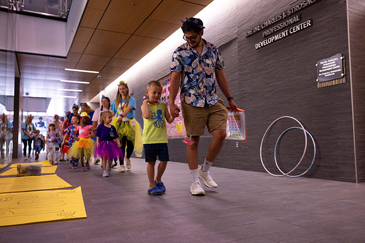 A croup of young children walk down a hallway, each holding hands with an adult