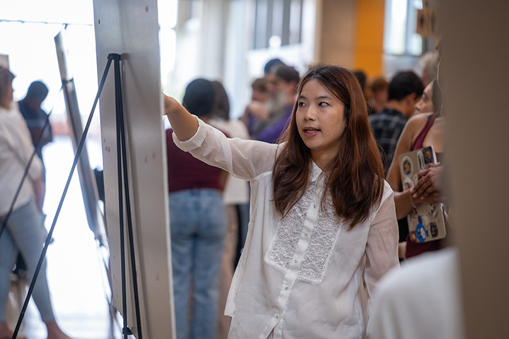 A female college student wearing a white shirt stands in front of a poster board, taking to a group of people
