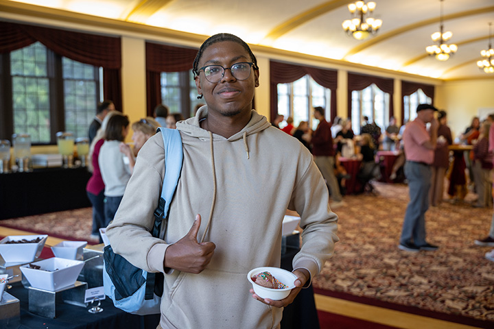 A CMU student with a backpack over their shoulder gives a thumbs up while holding a bowl of chocolate ice cream