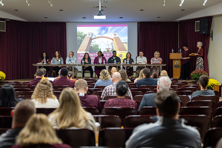 A panel of nine people sit at a table on a stage during the CMU Caring University event