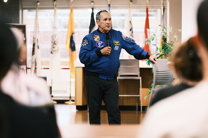 Astronaut John Herrington stands in front of a room with a microphone addressing the crowd in a blue jacket with honorary badges.