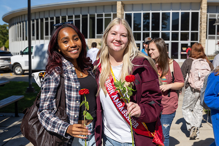 Two female college students hold roses and pose for a photo outside