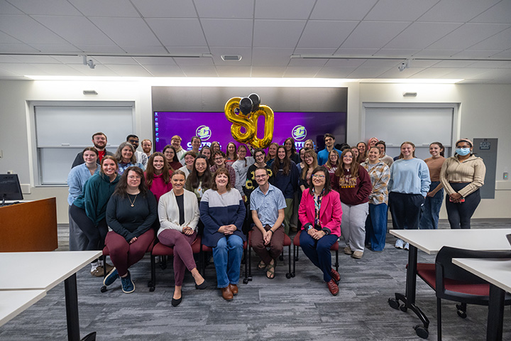 A group of two dozen people pose for a photo in a classroom celebrating 80 years of speech services and the audiology clinic