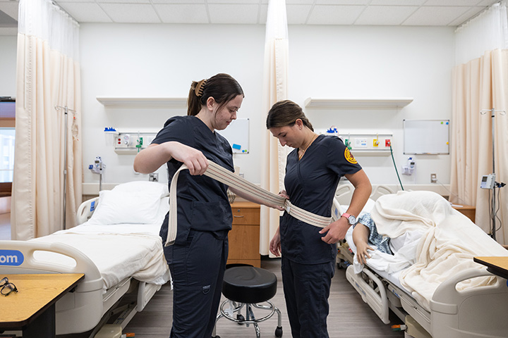 Two nursing students stand in a hospital room, one placing a very large belt on the other