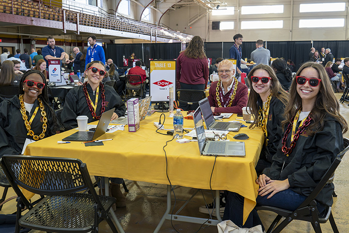 A group of five college students wearing sunglasses sit at a table covered in a yellow table cloth