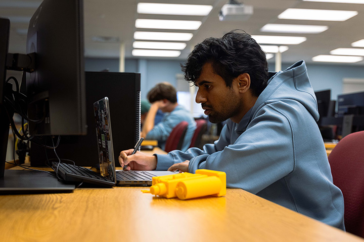 A student sits at a computer, holding a pen and writing on a piece of paper
