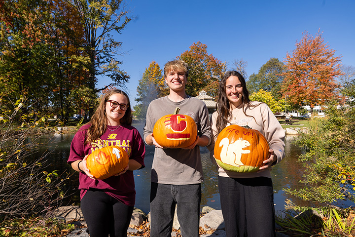 Three students stand outside holding painted pumpkins including one with a painted Action C
