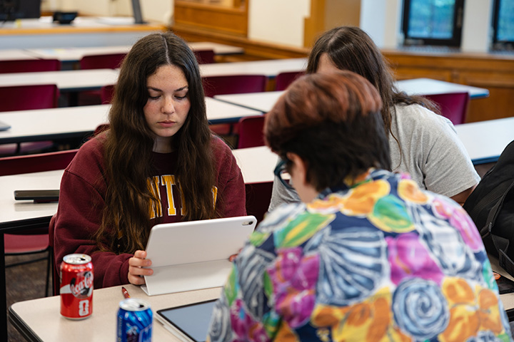 Three students sit at a table working on an iPad while cans of soda pop sit on the table.