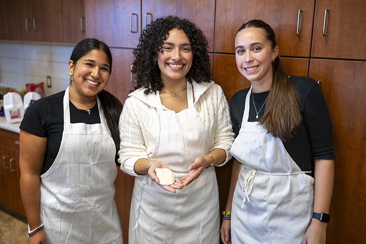 Three female college students wearing aprons pose for a photo inside a kitchen