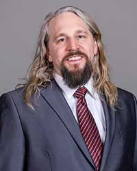 A man with long blond hair in a navy suit jacket and maroon tie smiles at the camera