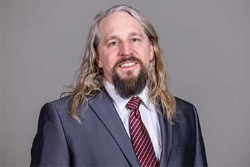 A man with long blond hair in a navy suit jacket and maroon tie smiles at the camera