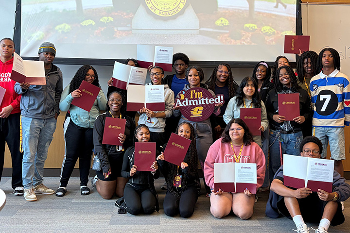 Inside a classroom, a group of high school students pose for a group photo while holding papers saying they have been admitted to CMU