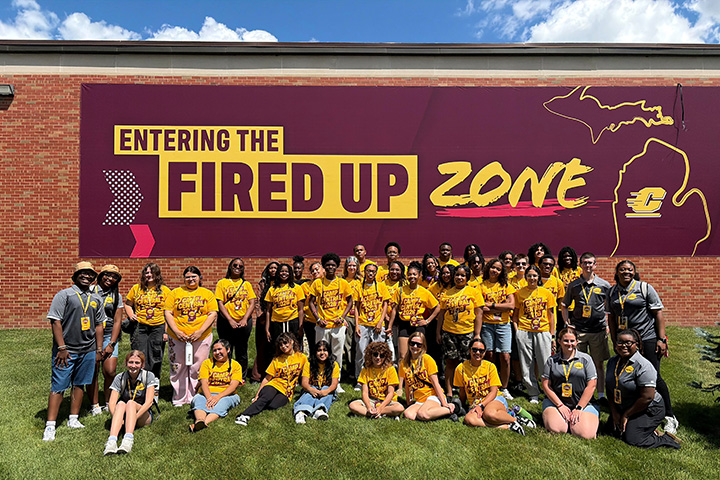 A large group of high school students pose for a photo in front of a building that has a large sign reading Entering The Fired Up Zone