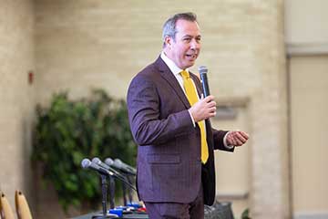 A man in a maroon suit and gold tie speaks into a microphone