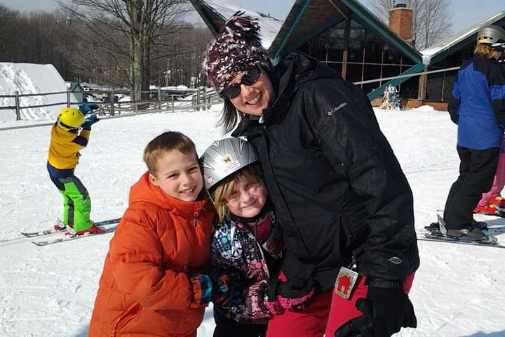 A woman in a winter hat and jacket poses with her two children on a ski slope