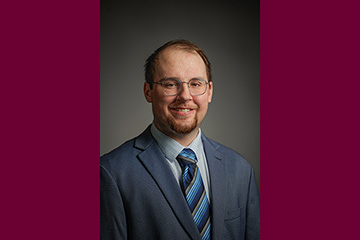 A man with glasses and a close-cropped beard and wearing a suit poses for a professional photograph against a brownish background.