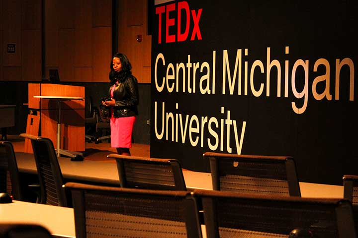 A Black woman in a pink skirt stands in front of a large screen that reads TEDxCentral Michigan University
