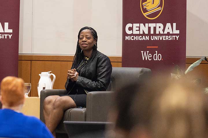 A Black woman in a black suit sits in a grey chair on a stage with the CMU logo behind her