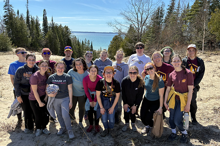 Nearly 20 CMU honors students pose for a photo on Beaver Island with Lake Michigan and trees in the background