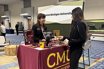 A woman stands behind a small table covered in a CMU tablecloth as another woman talks to her during a recruiting event