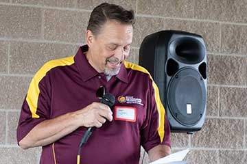 A man in a maroon and gold polo shirt stands next to a black audio speaker and speaks into a microphone.