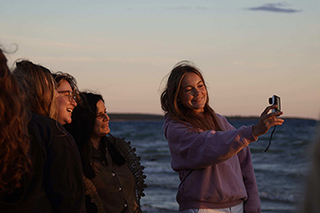 A student holds a camera to take a selfie of herself and a group of students while standing on the beach on Beaver Island