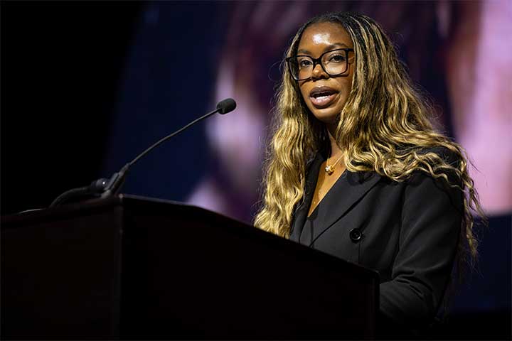 A young woman in a dark suit speaks into a microphone at a podium