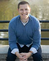 A smiling man in a blue sweater is sitting on a bench in front of a lake