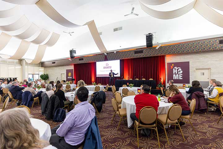 A room full of people seated at round tables watch as a man onstage speaks