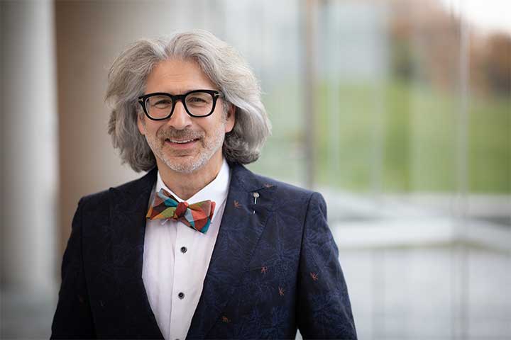 A man with long fluffy grey hair wearing glasses and a colorful bowtie smiles into the camera