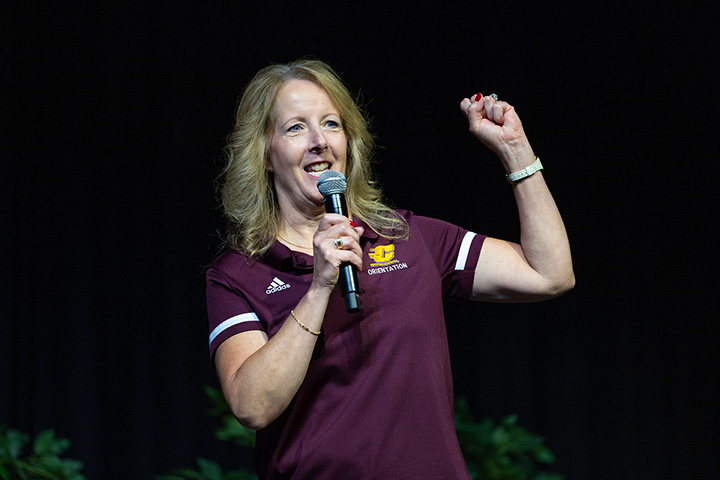 A woman in a maroon polo shirt holds a microphone in one hand while the other emphasizes the words she is saying